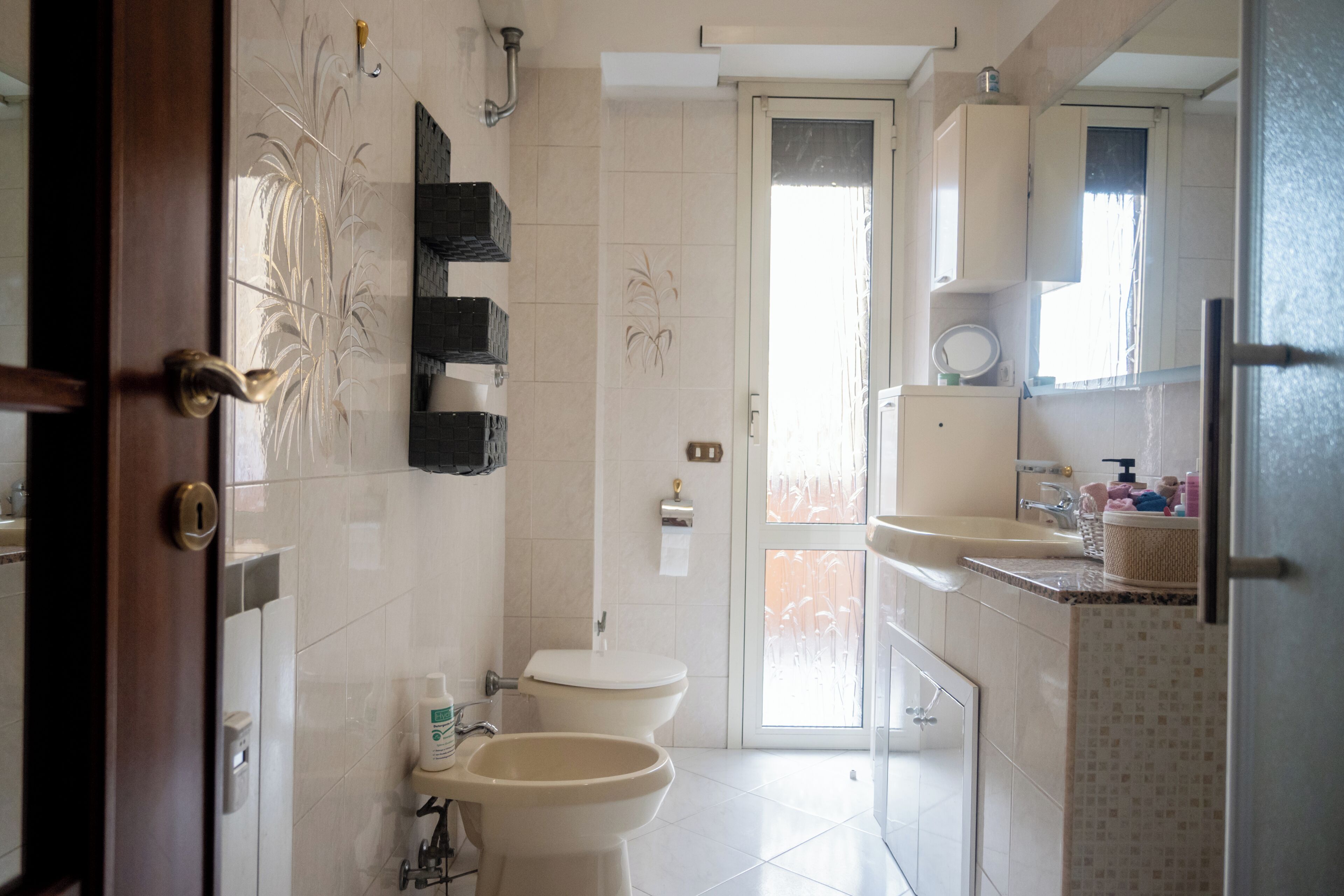 Une salle de bain comprenant des toilettes, un bidet et un lavabo, avec un sol carrelé et une porte donnant sur une autre pièce.