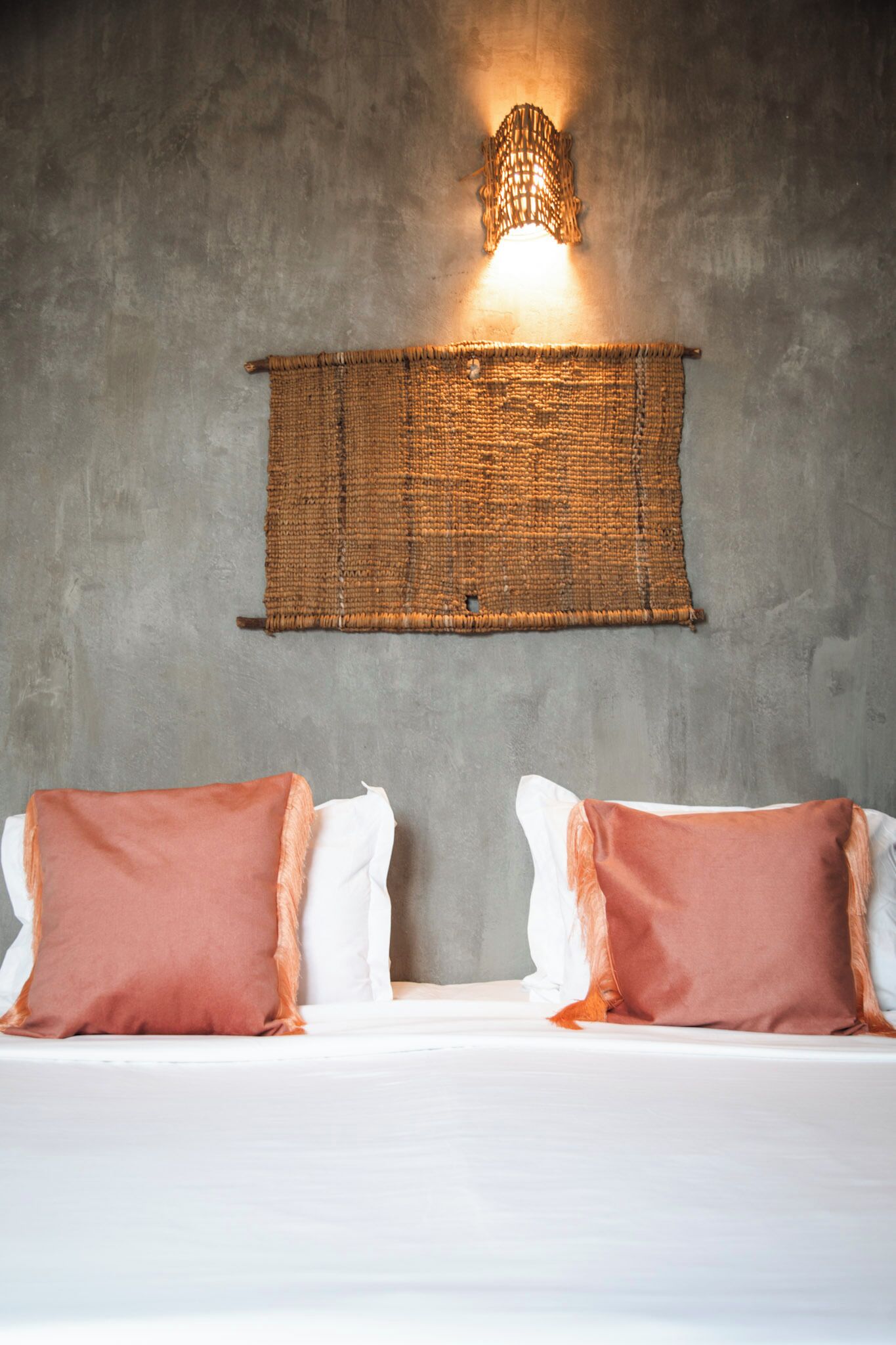 A bed with two pillows, a woven wall hanging, and a wall-mounted light fixture.