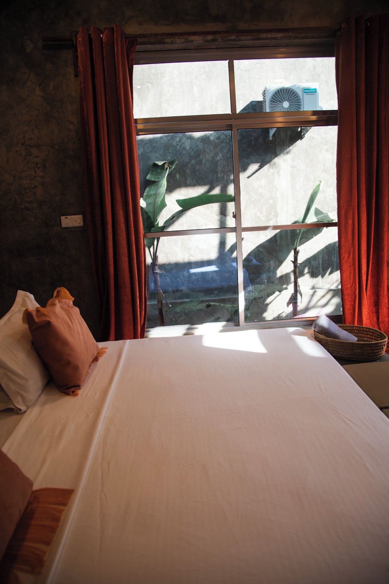 A room with a bed, a window with curtains, a wall-mounted air conditioner, and a plant outside.