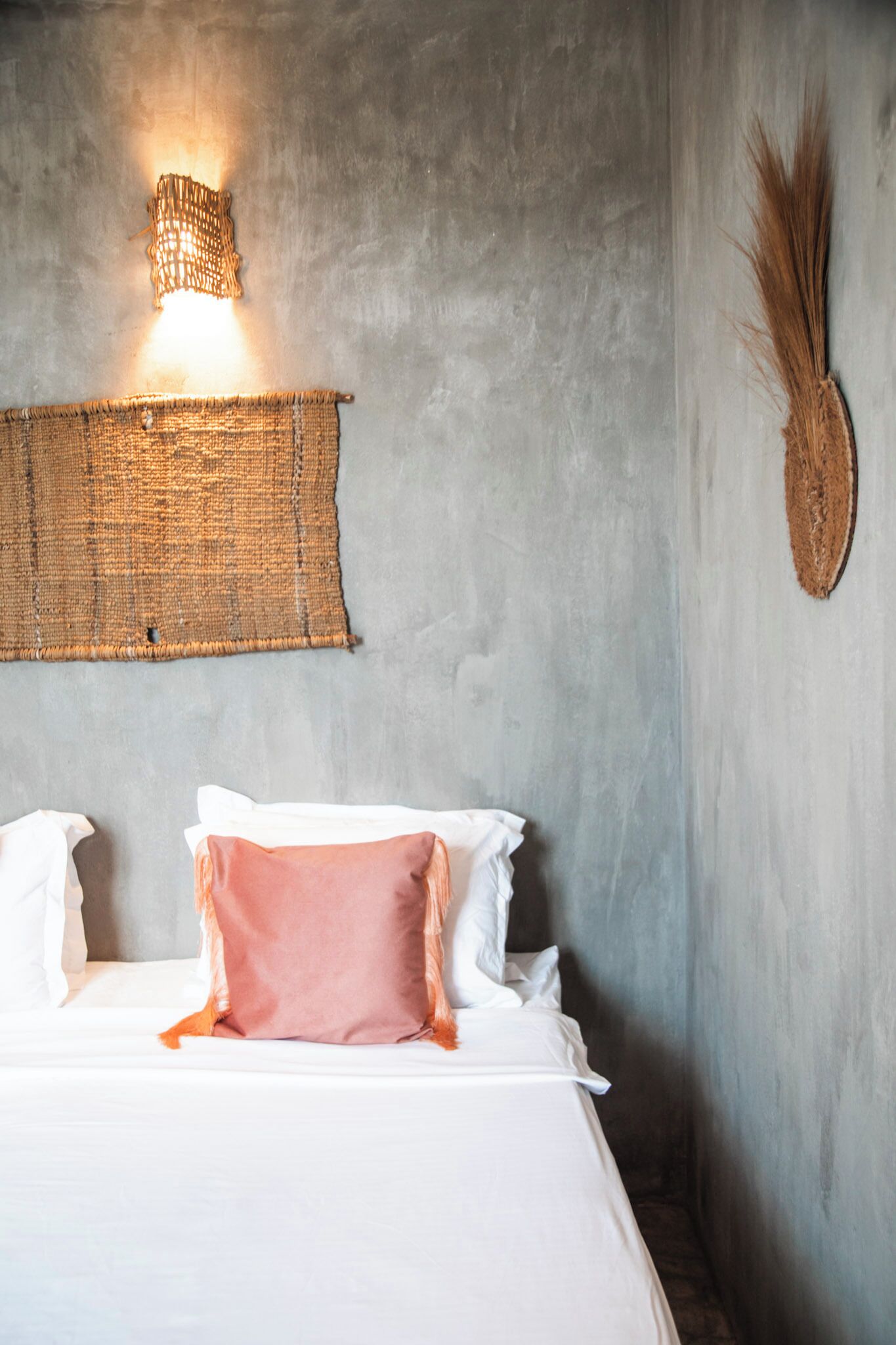 Uma cama com almofadas brancas e cor-de-rosa, um painel de parede tecido e uma luminária fixada na parede.
