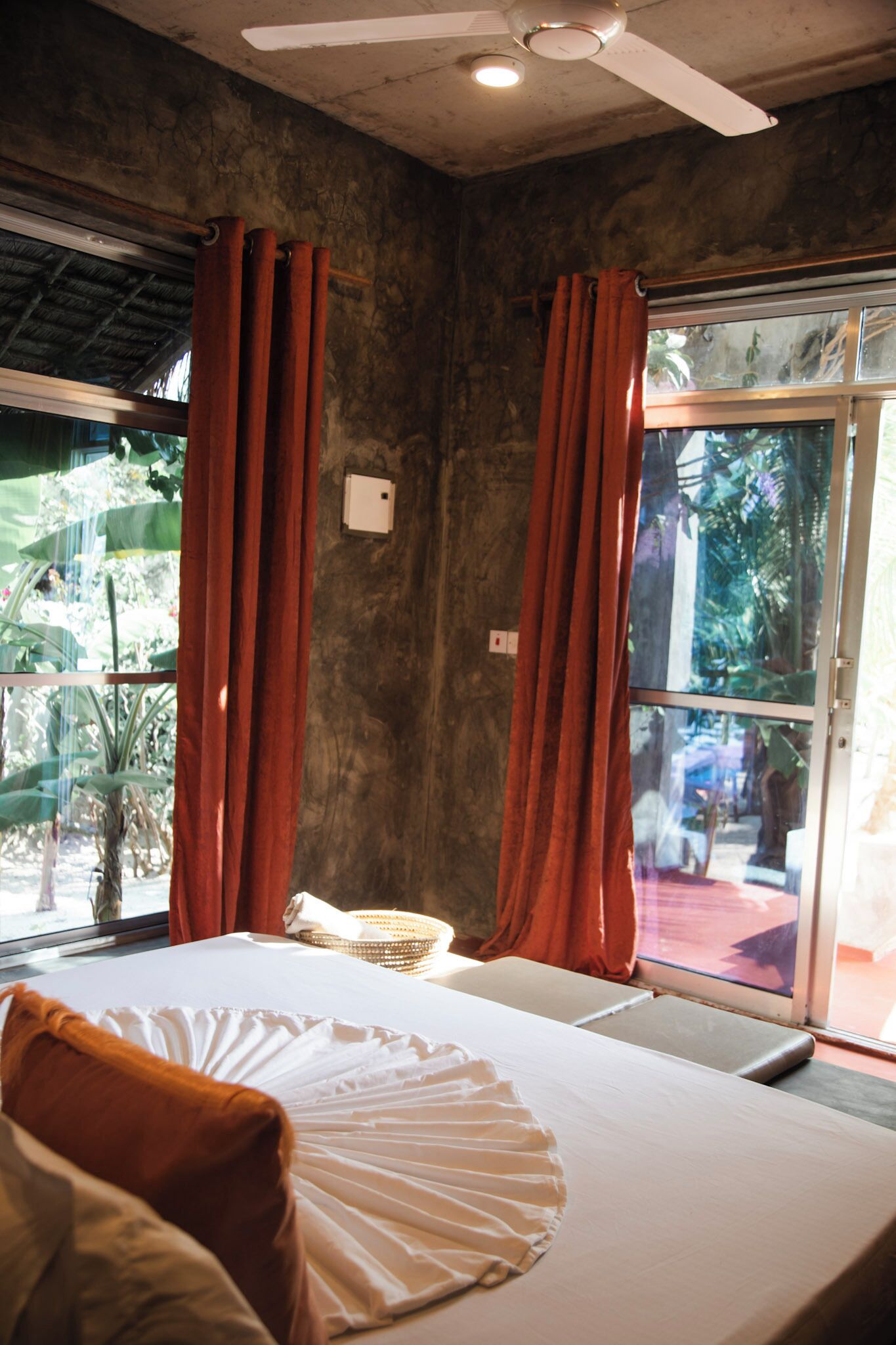 A room with a bed, curtains, a ceiling fan, and a window with a view of greenery.