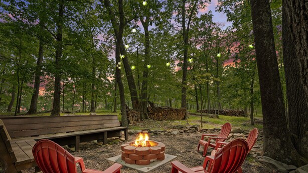 A fire pit surrounded by red Adirondack chairs in a forested area with a bench and a stone wall in the background.