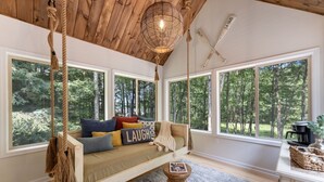 A cozy room with a hanging swing, large windows, and a view of the forest. - Lakeside Getaway Theatre, Indoor Pool and Game Room (Gouldsboro)
