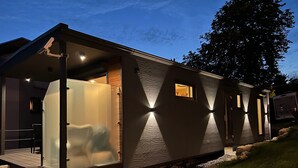 A modern RV with a covered deck and a small window, parked on a gravel driveway. - Gemütliches 4 Ferienhaus mit Whirlpool, Fitness, Infrarotsauna, Ebike, Alpakas (Braunfels)