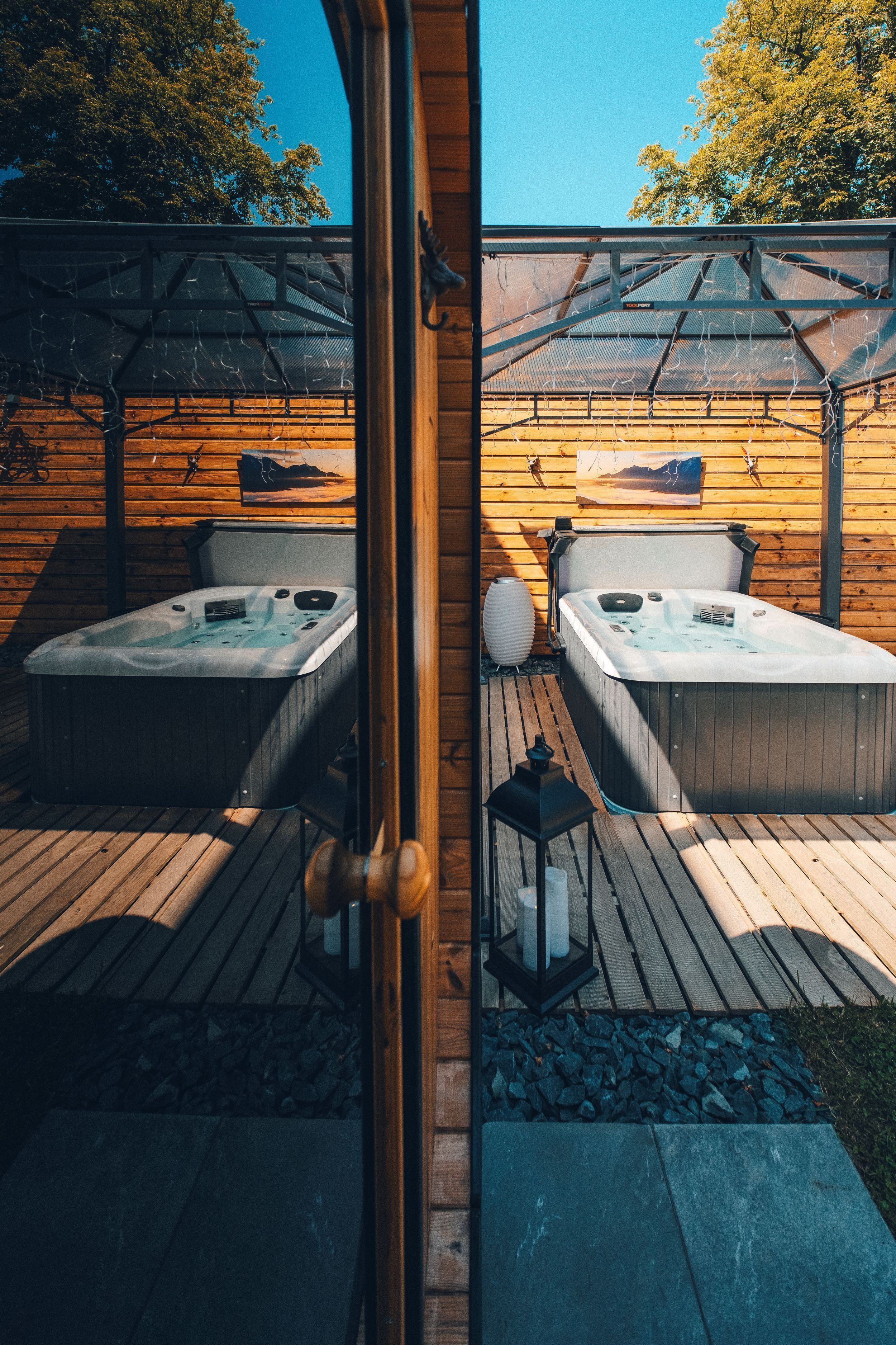 Eine Holzterrasse mit zwei Whirlpools unter einer Pergola, umgeben von Holzwänden und einem Steinrand.