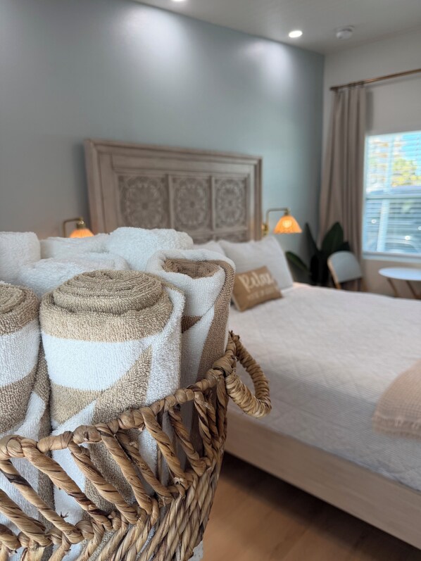 A neatly made bed with rolled towels in a woven basket, a headboard with a decorative panel, and a window with curtains.