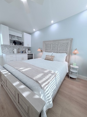 A bedroom with a bed, bedside lamps, a nightstand, a small table, and a kitchen area with cabinets and appliances. - Seaside Studio- POOL & Steps to the Beach!  (Madeira Beach)