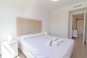 Ein ordentlich gemachtes Bett mit weißen Leinen und zwei gefalteten Handtüchern, ein Nachttisch mit Lampe sowie ein hölzernes Kopfteil.