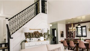 Ein Wohnzimmer mit einem beigen Sofa, einer Treppe mit schwarzen Geländern, einem Essbereich mit rosa Stühlen und einer Kristallleuchtre.