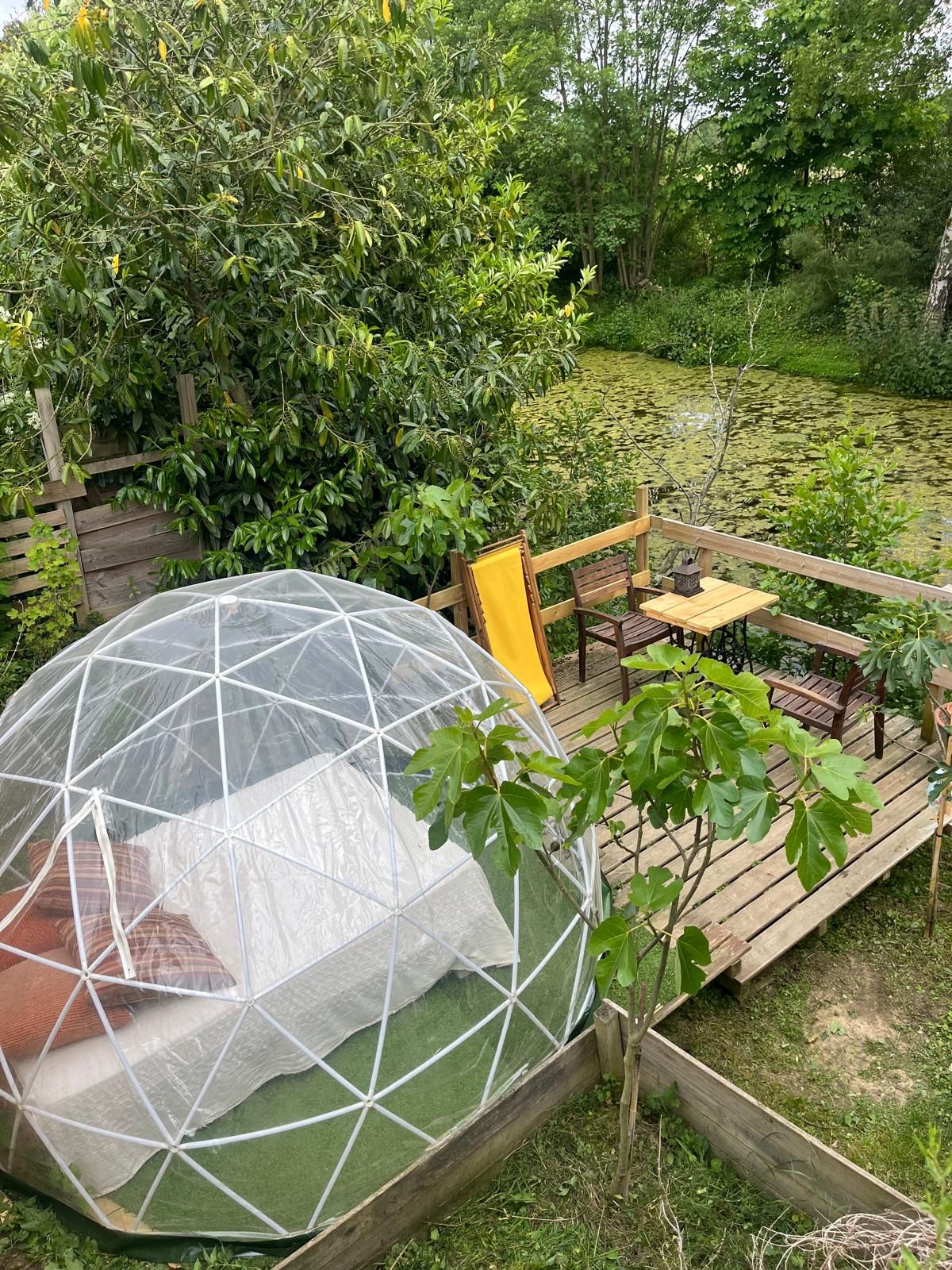A greenhouse structure with a transparent dome, a wooden deck with seating, and a surrounding garden.