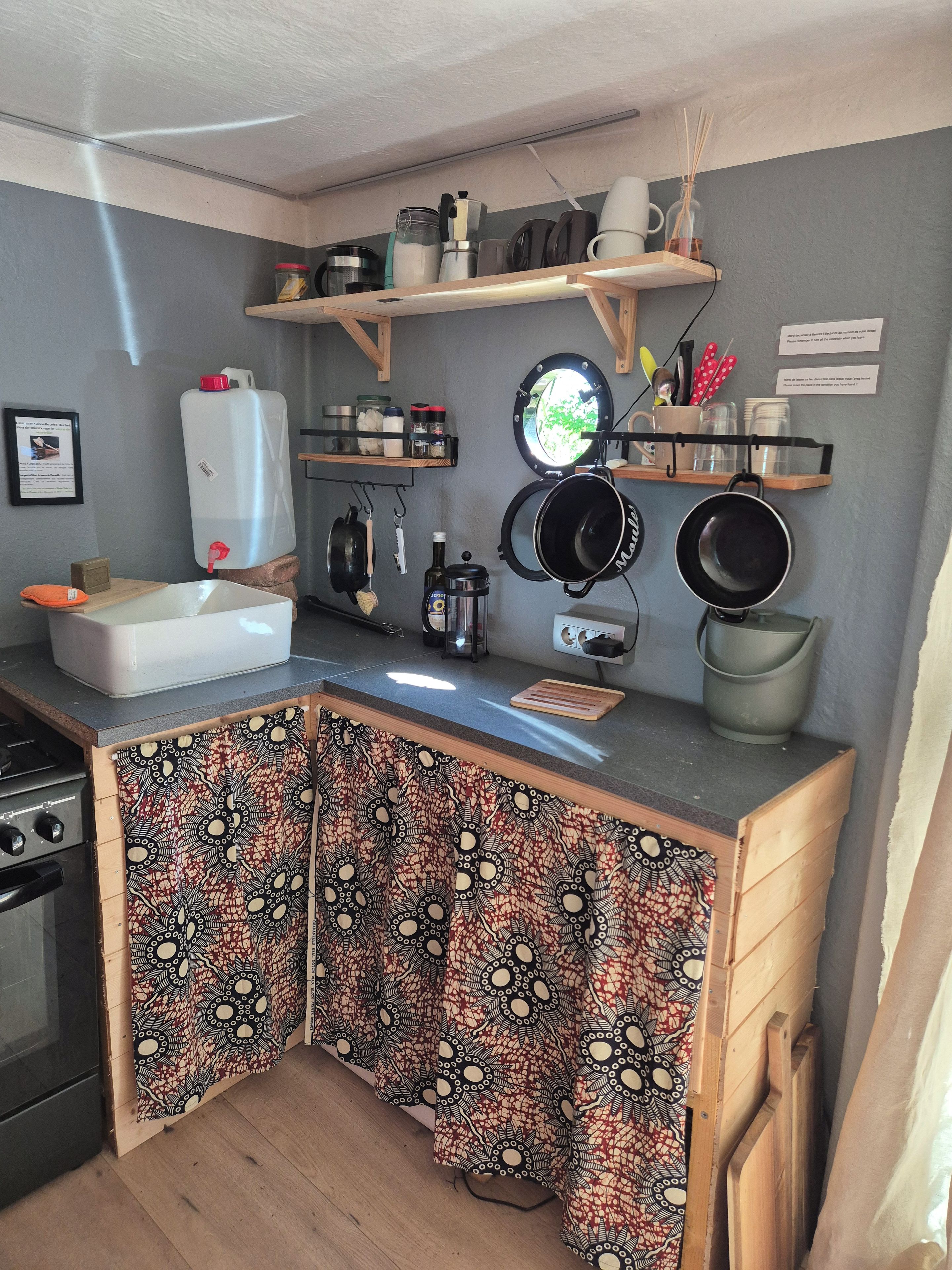 Una cocina compacta con un lavamanos, estufa y varios utensilios. Hay repisas sobre la encimera con frascos y una tetera.