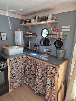 A compact kitchen with a sink, stove, and various utensils. There are shelves above the counter holding jars and a kettle.