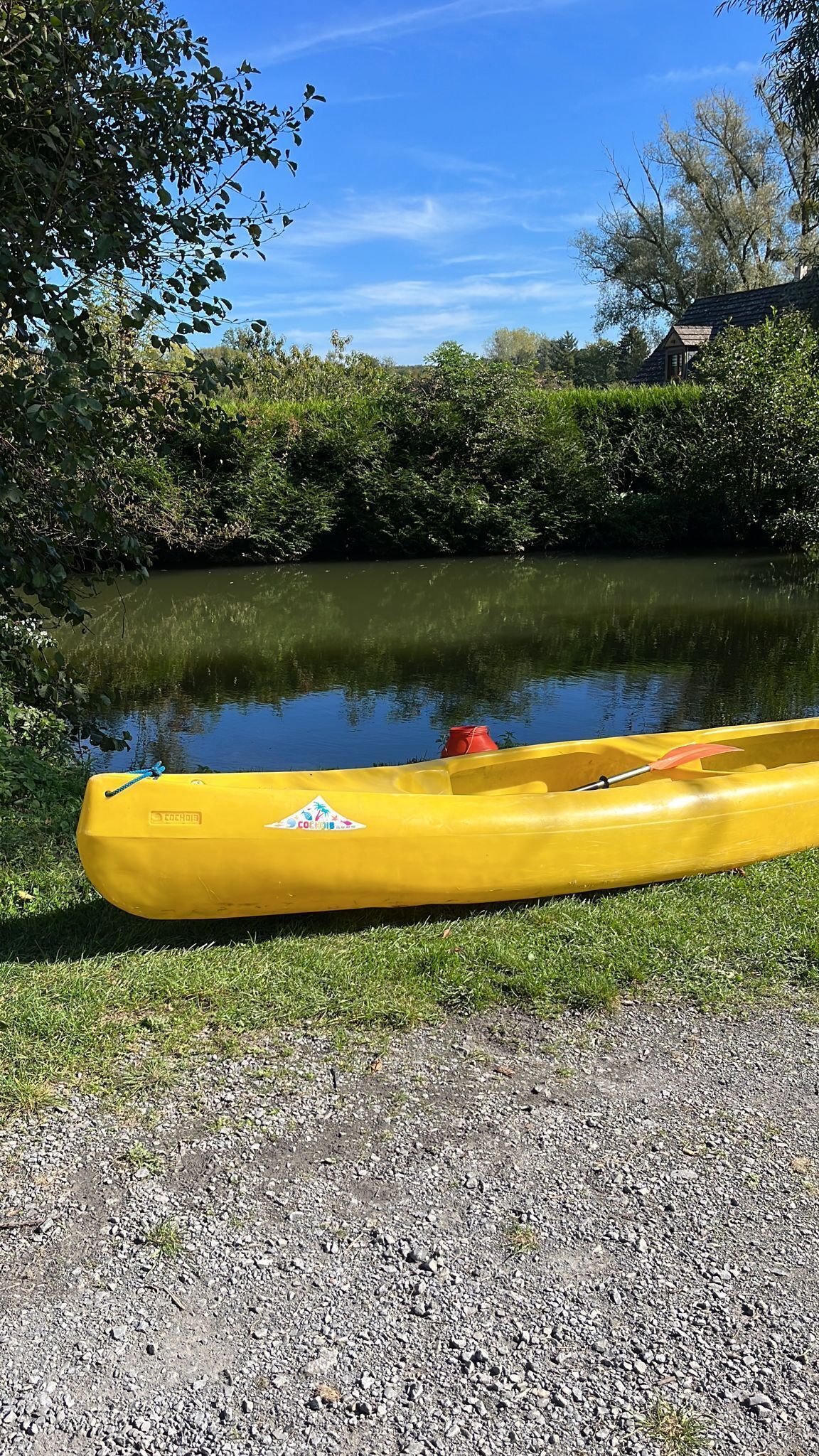 A yellow kayak with a red paddle and a COACH logo, resting on a gravel path beside a calm river.