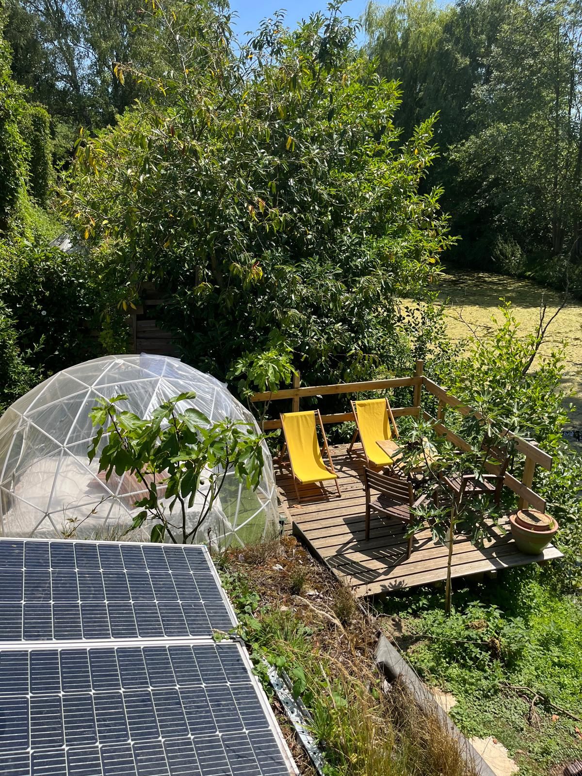 Una estructura tipo invernadero con una cúpula transparente, una terraza de madera con sillas amarillas y paneles solares.