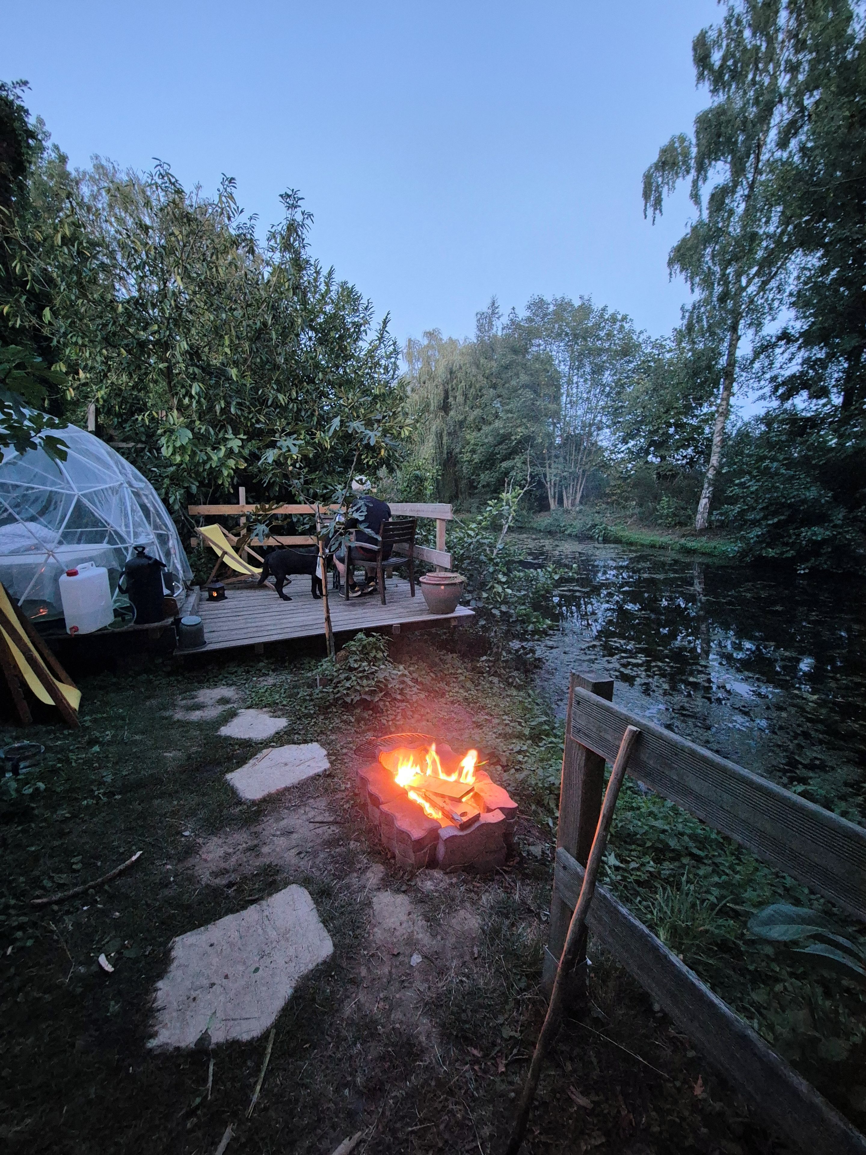 Un terrain de camping équipé d’un foyer, d’un espace abrité et d’une terrasse en bois donnant sur un plan d’eau.