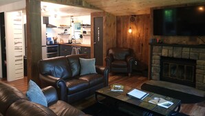 A cozy living room with a leather sofa, a coffee table, and a fireplace. There is a kitchen in the background with wooden cabinets and a sink. - POND IN THE PINES CABIN -COMPLETELY RENOVATED ON 27 ACRES - POND-HOCKING HILLS (McArthur)