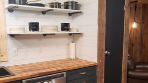 A kitchen with a wooden countertop, stainless steel dishwasher, and black cabinets. There are shelves above the counter holding pots and plates. A door and a couch are visible in the background. - POND IN THE PINES CABIN -COMPLETELY RENOVATED ON 27 ACRES - POND-HOCKING HILLS (McArthur)