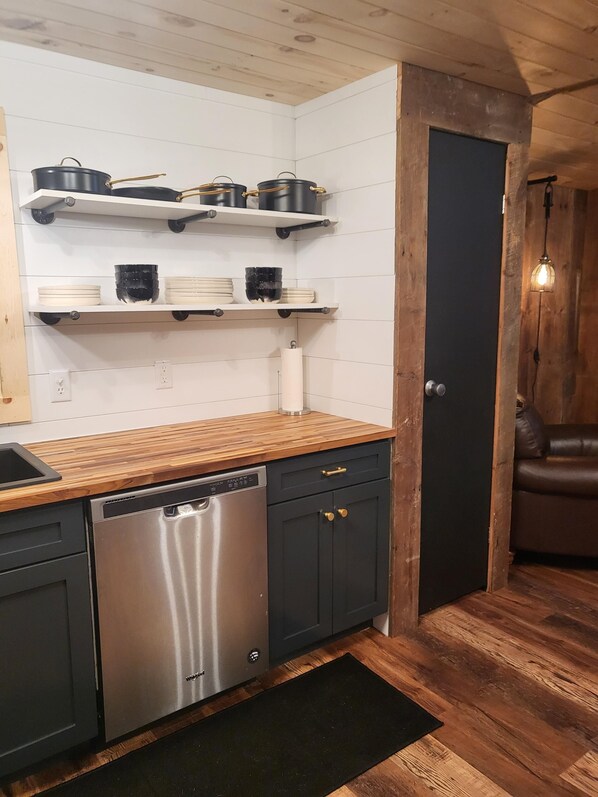 Une cuisine avec un plan de travail en bois, un lave-vaisselle en acier inoxydable et des meubles noirs. Des étagères au-dessus du plan de travail servent à ranger les casseroles et les assiettes. On aperçoit une porte et un canapé à l’arrière-plan.