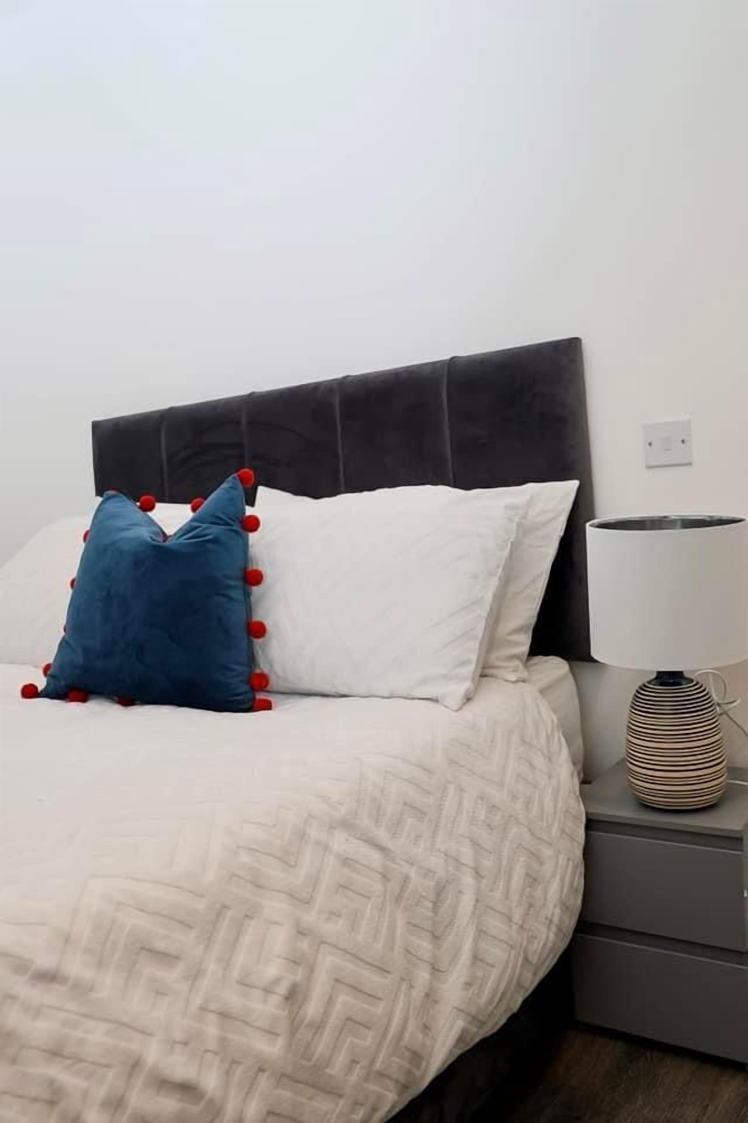 Uma cama com colcha estampada, duas almofadas, um abajur e uma mesa de cabeceira cinza.