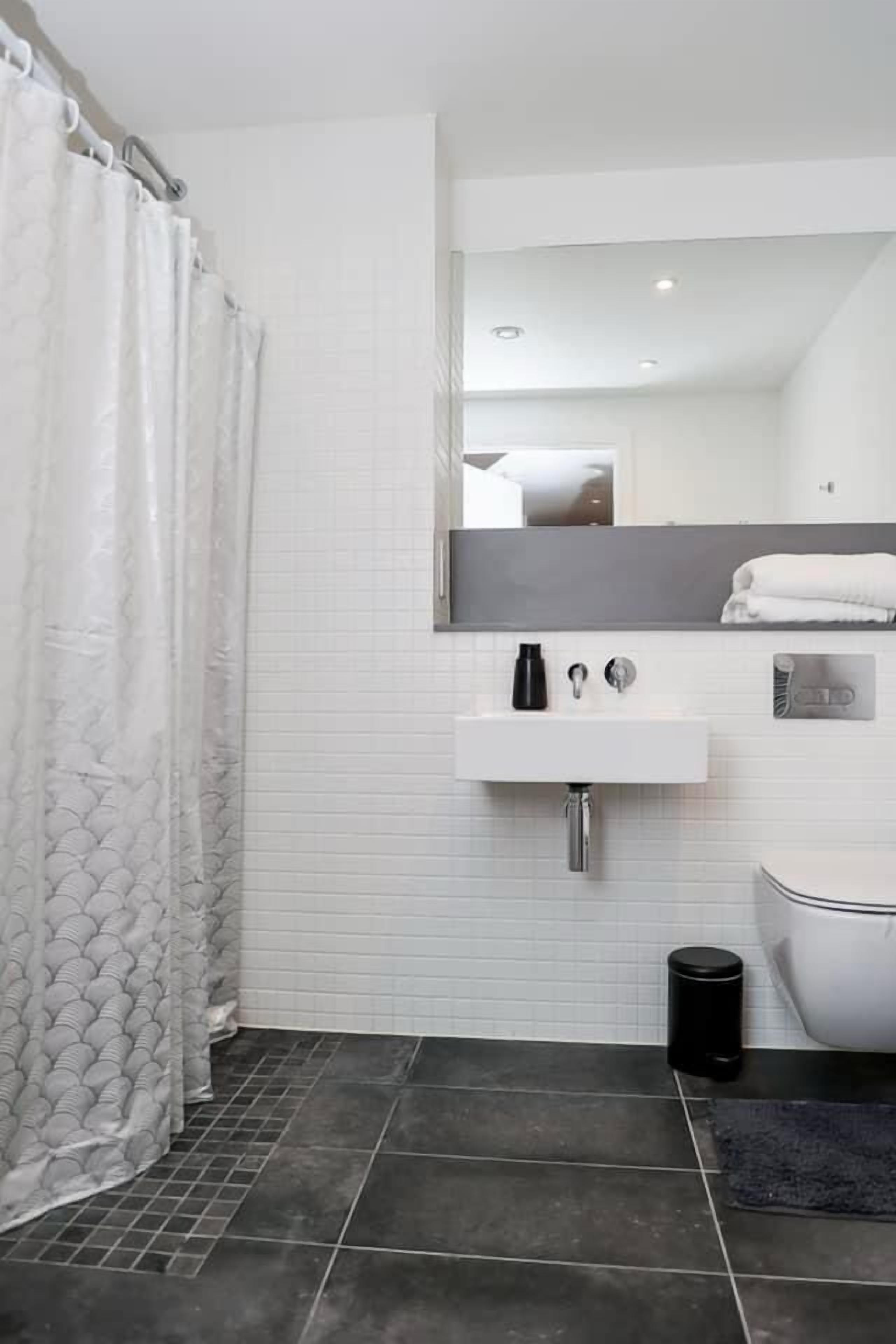 A modern bathroom with a white tiled wall, a black trash bin, a white sink, and a toilet.