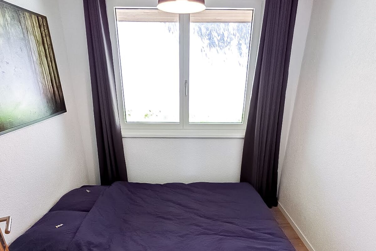 Une petite chambre avec un lit simple, une fenêtre, un plancher en bois et un luminaire au plafond.