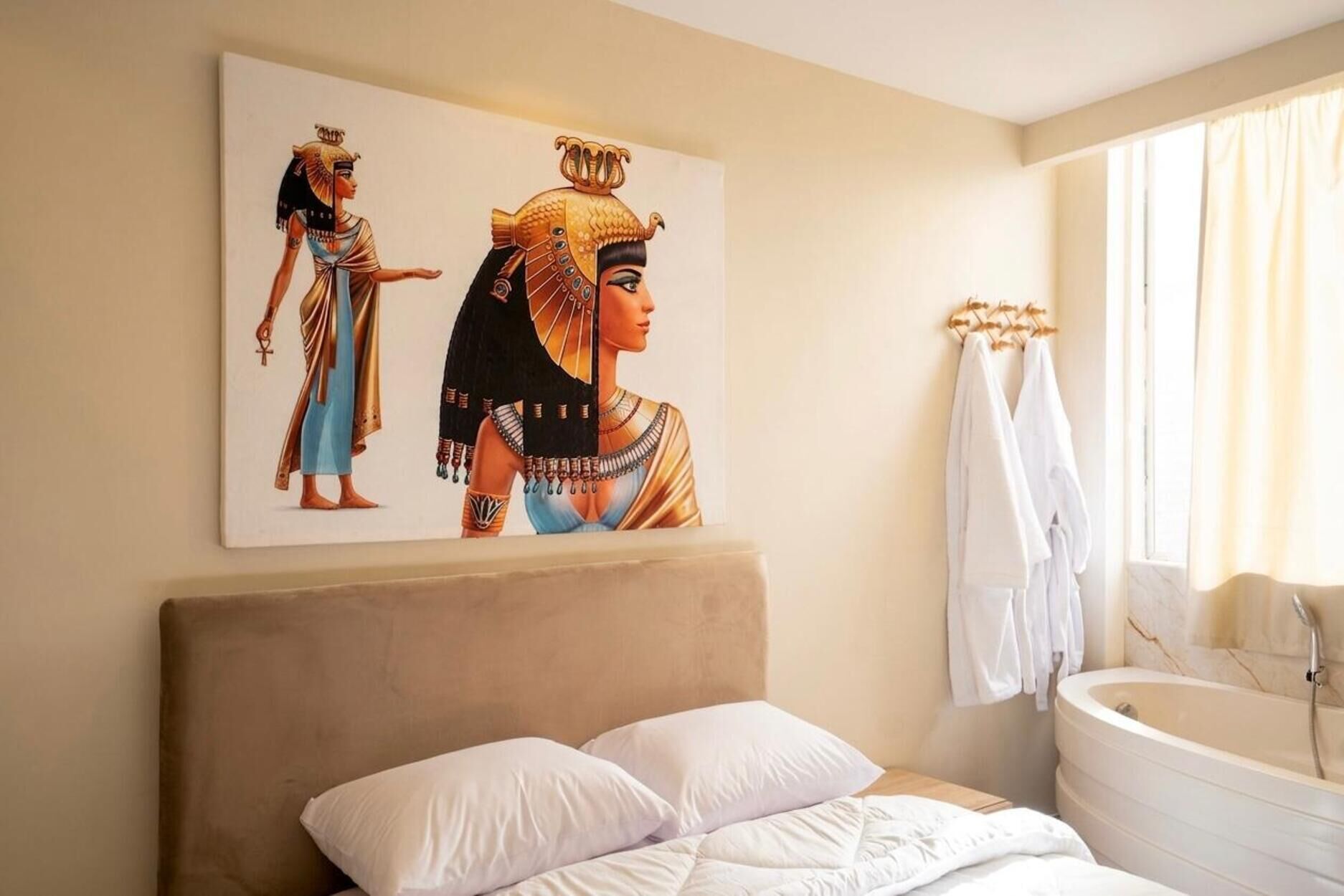A bedroom with a bed, pillows, a wall-mounted painting of Egyptian figures, a window with curtains, and a bathtub.