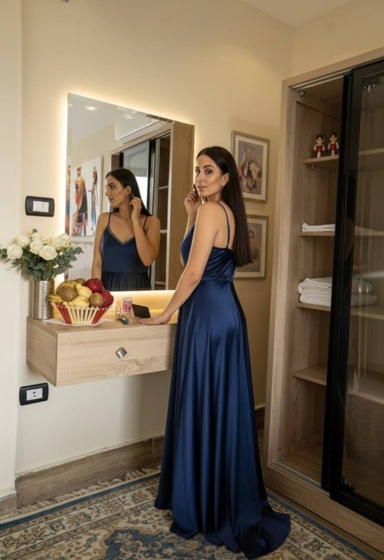 Una mujer con un vestido azul marino largo está de pie en una habitación con un tocador de madera, un espejo y un frutero pequeño en la encimera.