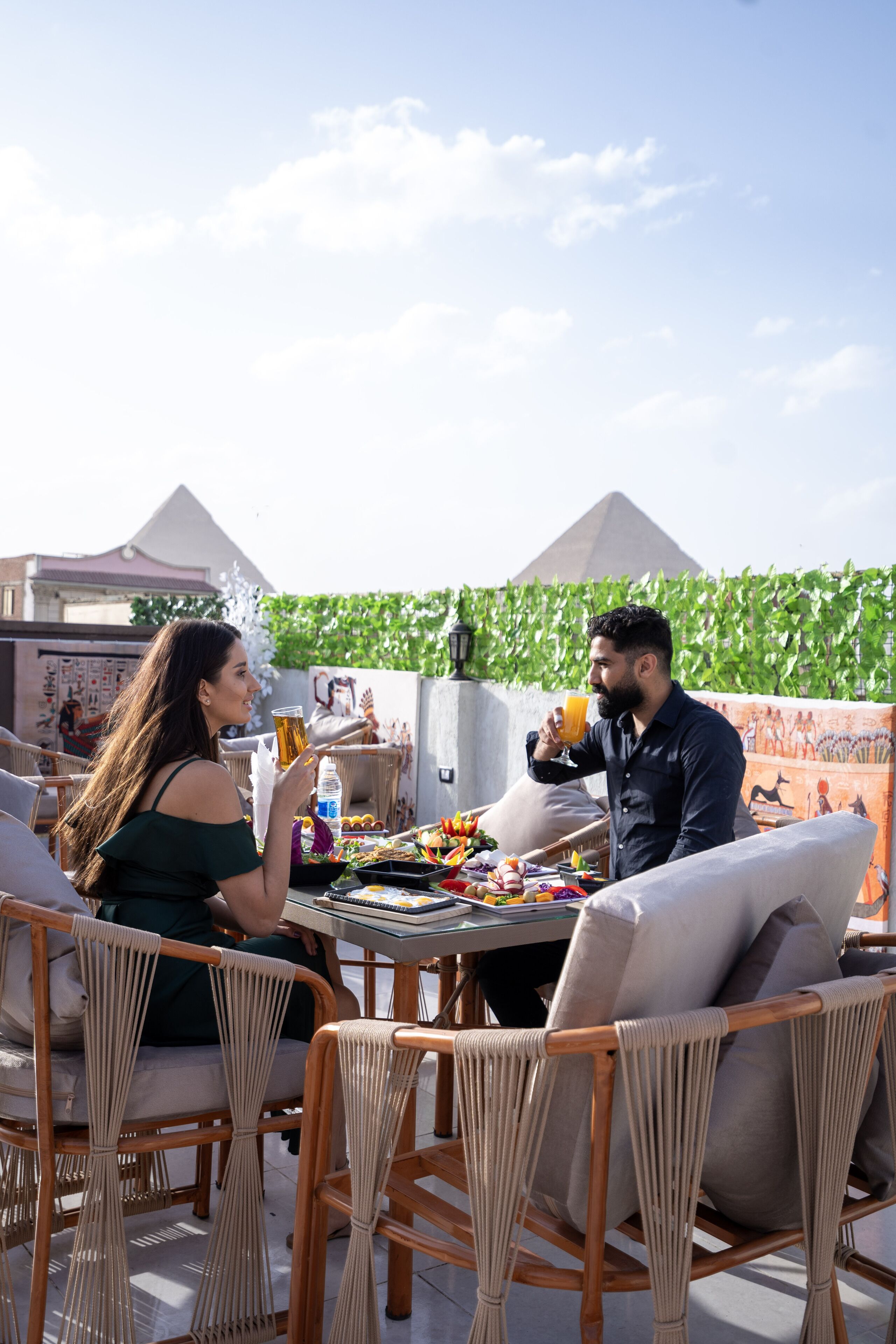 A couple dining at an outdoor table with wicker chairs, a view of the pyramids, and a green leafy backdrop.