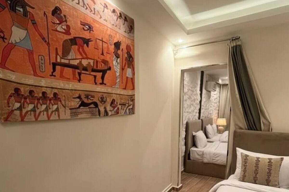 A hotel room with a large wall mural depicting ancient Egyptian scenes, a bed with white linens and patterned pillows, a wooden floor, and a small woven rug.