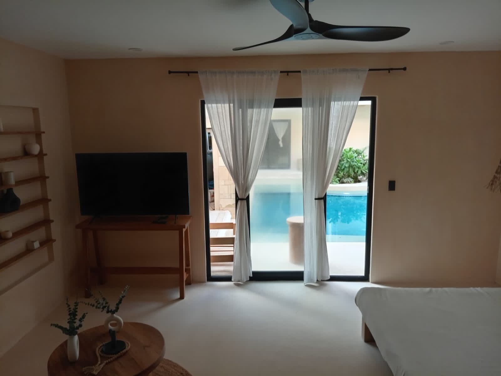 Quarto de hotel com televisão de tela plana em suporte de madeira, cama, ventilador de teto e vista para uma piscina através de uma porta de vidro.