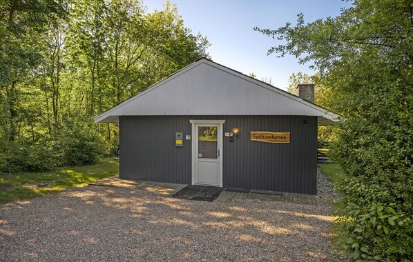 A small, single-story building with a grey exterior and a white door, surrounded by lush greenery. - Beautiful home in Toftlund with WiFi (Toftlund)