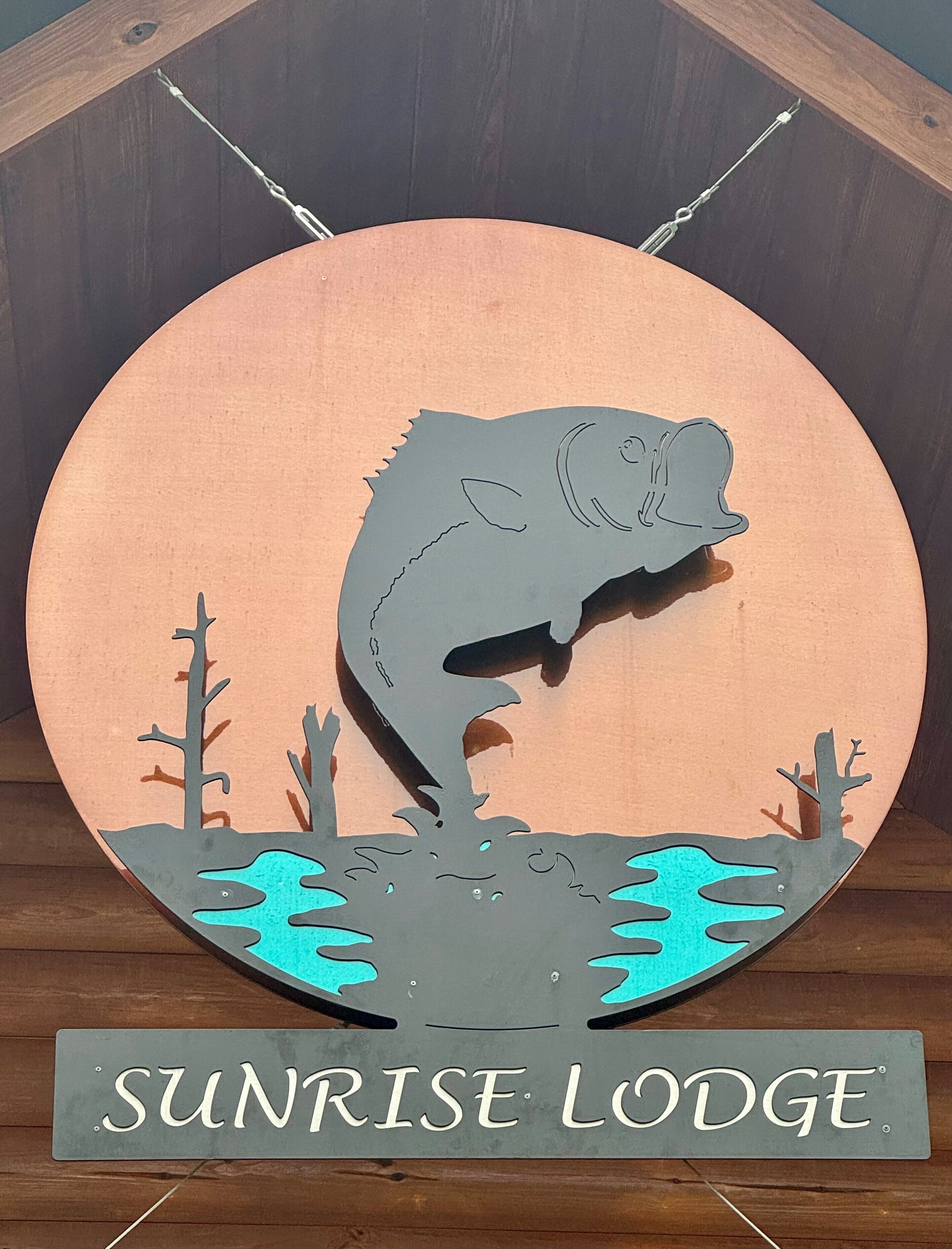 Ein rundes Holzschild mit einer Silhouette eines springenden Fisches und Bäumen, beschriftet mit 'SUNRISE LODGE'.