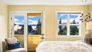Un dormitorio con cama, una silla, una lámpara y una ventana con vistas a árboles y un edificio.