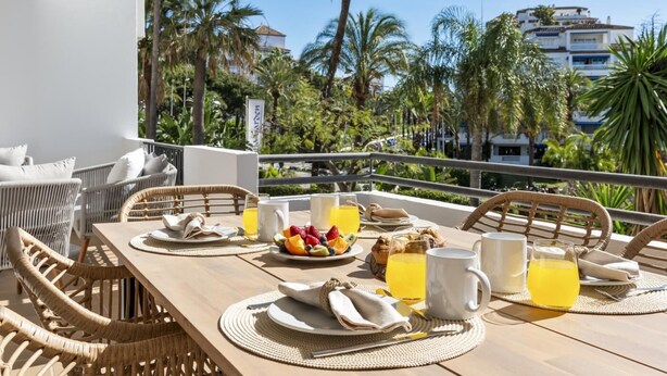 A dining table set with plates, cutlery, and drinks, overlooking a tropical landscape with palm trees and buildings.