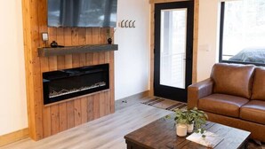 Ein Wohnzimmer mit Holzboden, einem an der Wand befestigten Flachbildfernseher, einem Kamin, einer Couch und einem Couchtisch mit Pflanze.