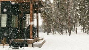 Eine Hütte mit Veranda und überdachter Sitzgelegenheit, umgeben von hohen Kiefern und bedeckt mit Schnee.