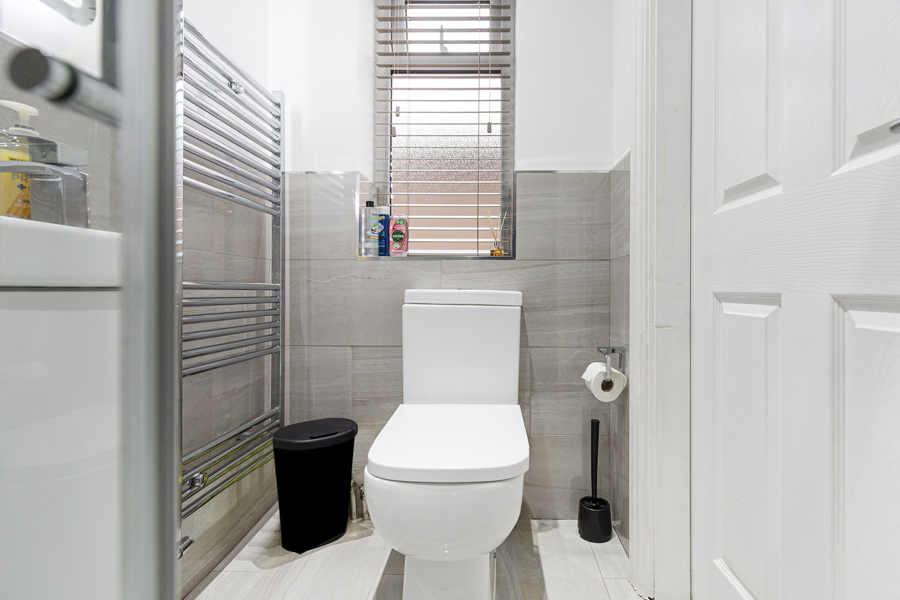 Een moderne badkamer met toilet, een raam met zonwering, een prullenbak en een toiletrolhouder.