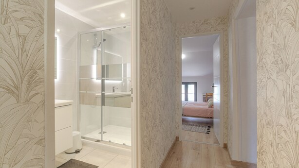 A modern bathroom with a glass shower enclosure, a toilet, and a sink. A hallway leads to a bedroom with a bed and a nightstand. - Gîte La Terrasse, near CNPE and Via Rhôna, ideal for cycling enthusiasts! (Lagnieu)