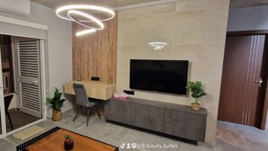 A modern living room with a flat-screen TV, a wooden cabinet, a dining area with a table and chairs, and a sliding glass door leading to a balcony.