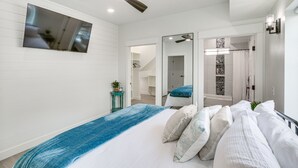 Ein modernes Schlafzimmer mit einem großen Bett, einem Deckenventilator, einem an der Wand montierten Fernseher und einem durchgehenden Türspalt einsehbarer Badezimmers.