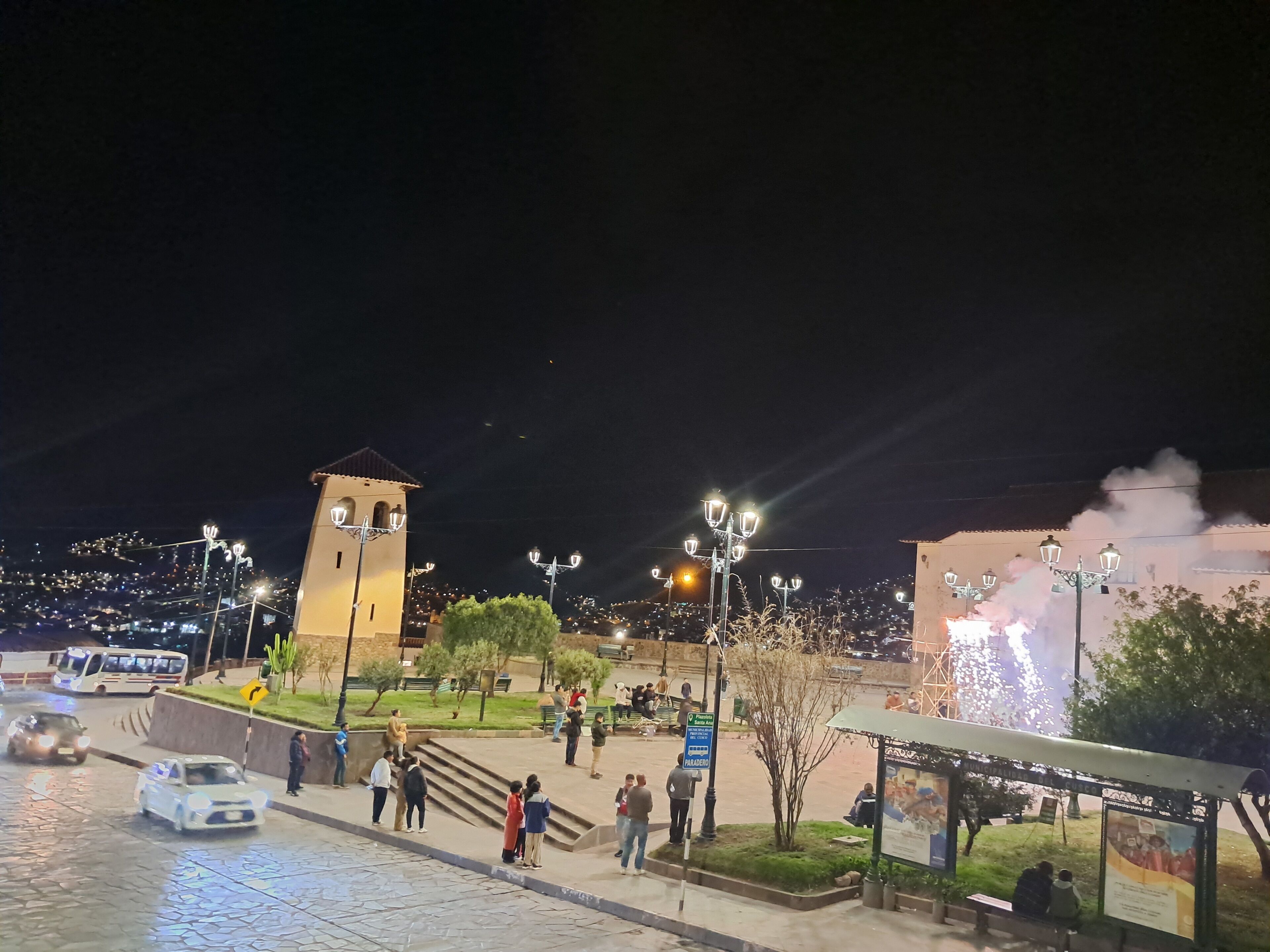 A nighttime urban scene with a prominent tower, street lamps, and a bus stop.