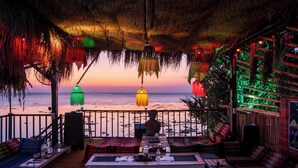 Un salón junto a la playa con vista al océano, techo de paja y una mesa lista para comer.