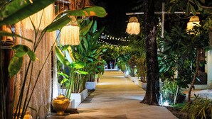 Un sendero bien iluminado bordeado de plantas en macetas y luces colgantes, que conduce a la entrada de un edificio.