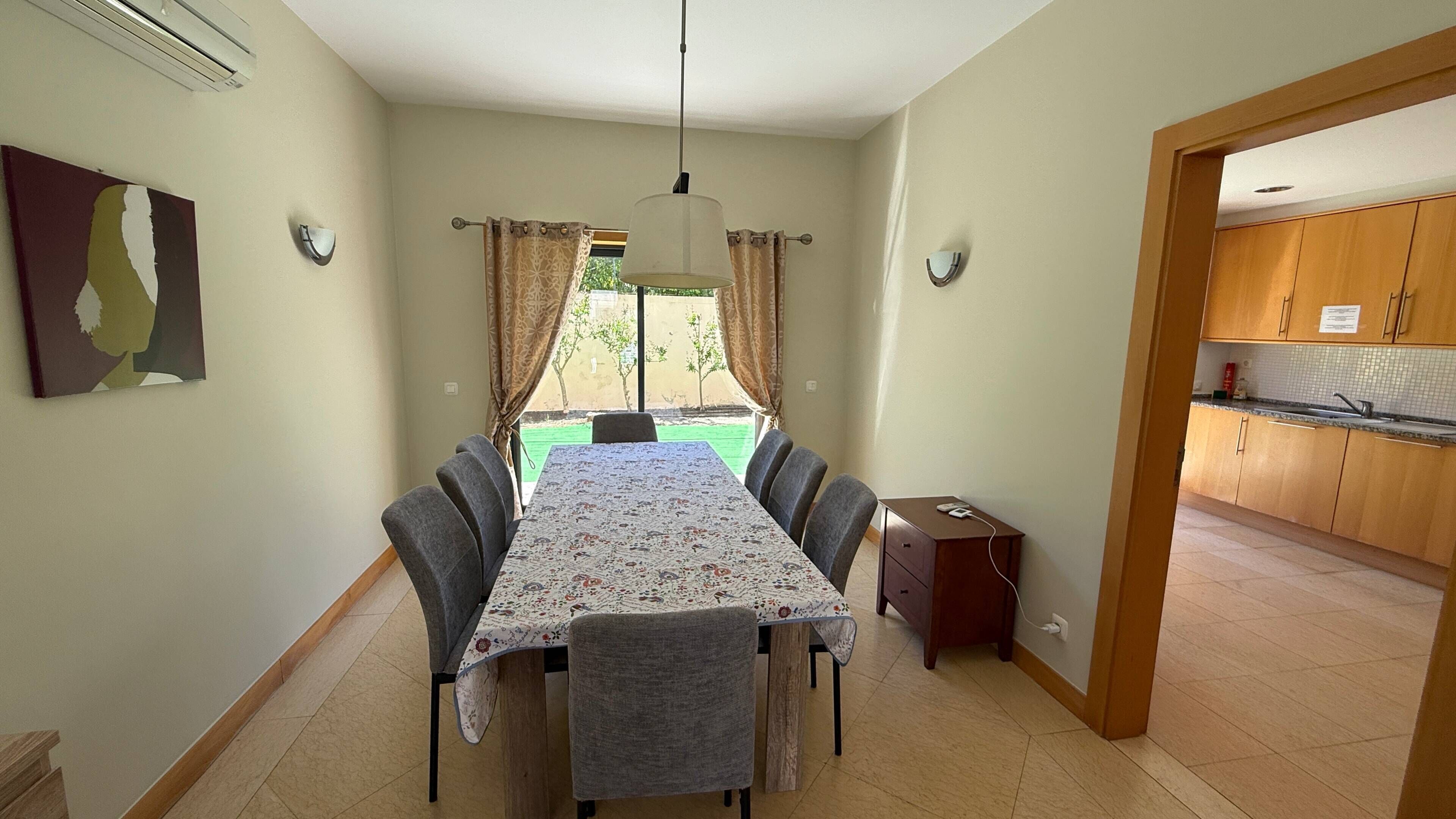 Un coin repas avec une table et des chaises, un luminaire suspendu, une vue par la fenêtre et une cuisine visible à travers une porte ouverte.