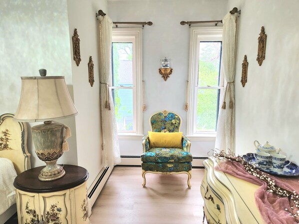 Une pièce comprenant un fauteuil classique, une table avec une lampe et une commode sur laquelle est posée une assiette remplie de tasses à thé.