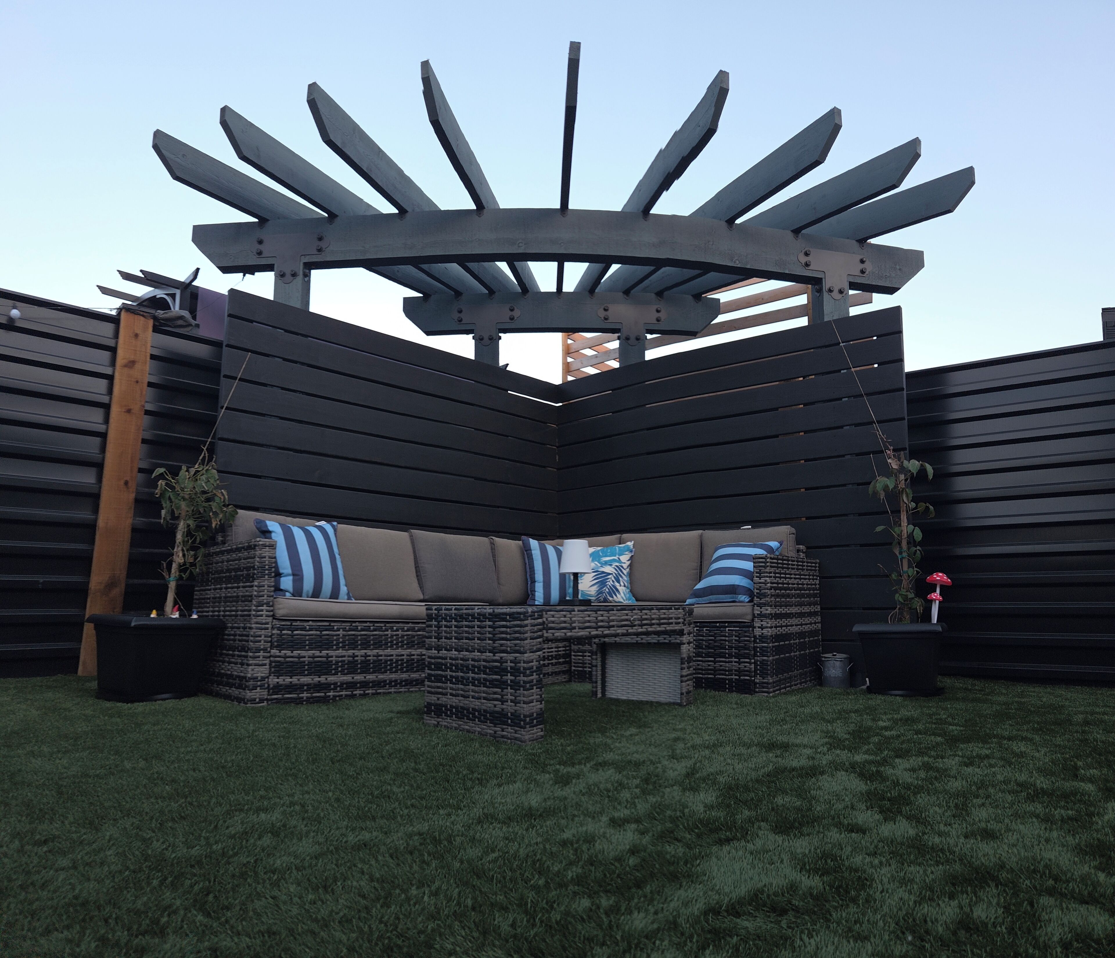 Un petit jardin aménagé dans un coin, avec une pergola en métal noir, un canapé en osier agrémenté de coussins et deux plantes en pot.