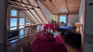 A cozy attic bedroom with a sloped ceiling, wooden beams, a ceiling fan, a bed, a nightstand, a lamp, a window, and a balcony railing with lights. - Experience Bethel charm near Sunday River  (Bethel)
