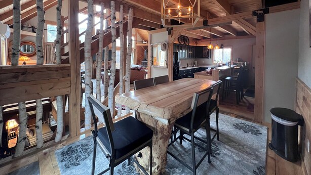 A rustic wooden interior with a dining area featuring a large wooden table and chairs, a kitchen with dark cabinets, and a staircase with birch logs. - Experience Bethel charm near Sunday River  (Bethel)
