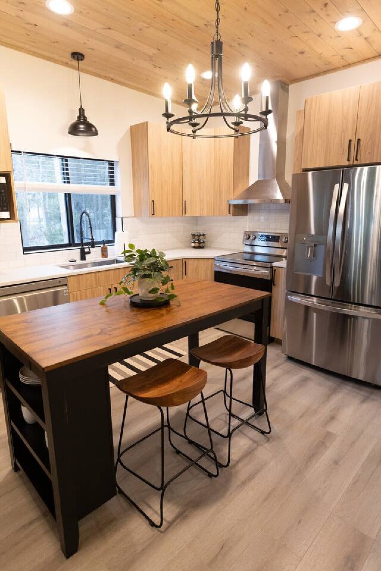 A modern kitchen with wooden cabinets, a stainless steel refrigerator, a central island with stools, and a chandelier.