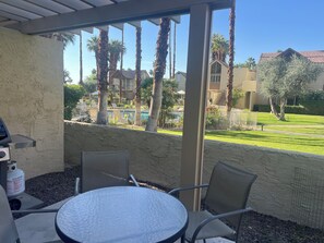 A covered patio with a round table and two chairs, overlooking a pool and residential buildings.